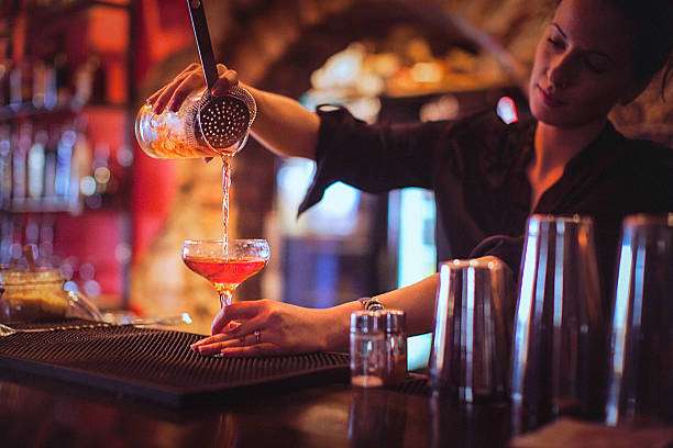 Bartender pouring a colorful drink through a strainer into a glass; a cocktail shaker is nearby.
