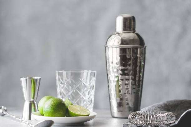Shiny metal cocktail shaker, jigger, limes, and glass on a grey background, ready for drinks.