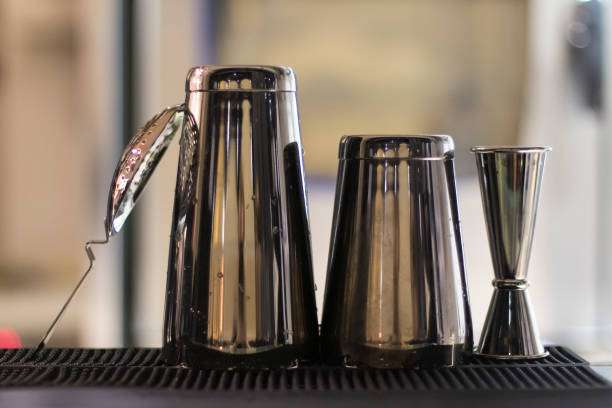 Metal cocktail shaker set including jigger and shaker cups on a dark bar mat.