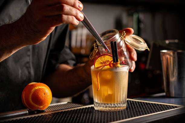 Bartender garnishes a cocktail with orange, while a Cocktail Shaker waits nearby.