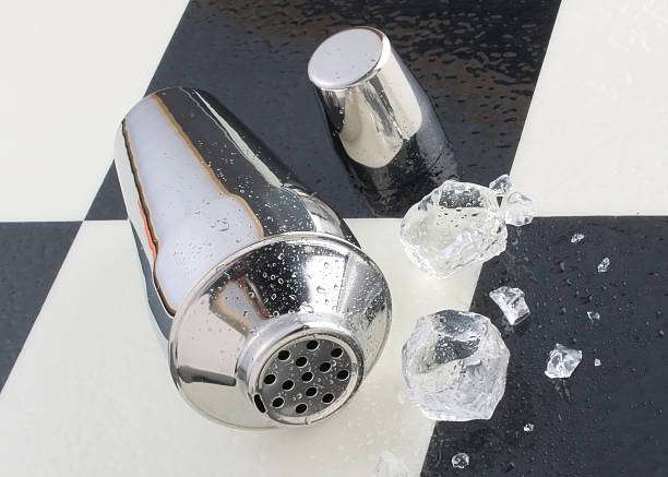 Metal cocktail shaker and ice cubes on a wet black and white tiled surface.